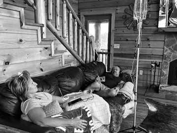 Black and white photo of a couple resting on a leather sofa in a log cabin living room, each on an IV drip