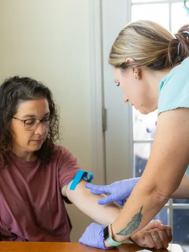 Meghan, in teal scrubs, starting an IV for a client seated at her kitchen table