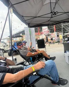 Men relaxing in reclining chairs under a canopy tent with IV drips at a race track, one giving a thumbs up